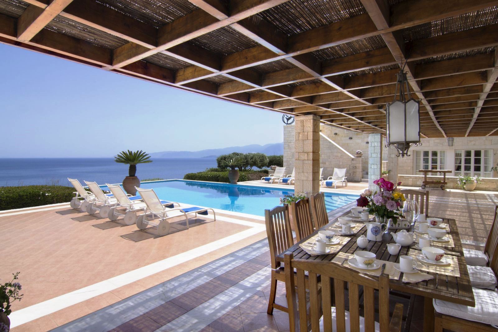 Covered terrace area with wooden table set for breakfast and chairs by the side of the pool at Villa Selene in Crete