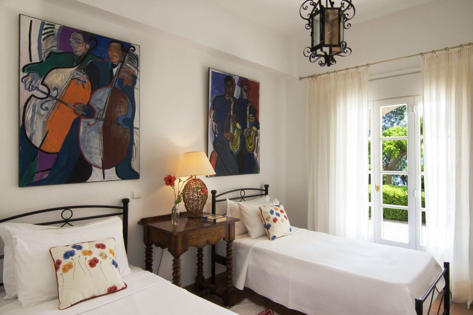 Twin beds with a wooden table between them and big pictures on the wall and a glass door out onto the garden at Villa Selene in Crete