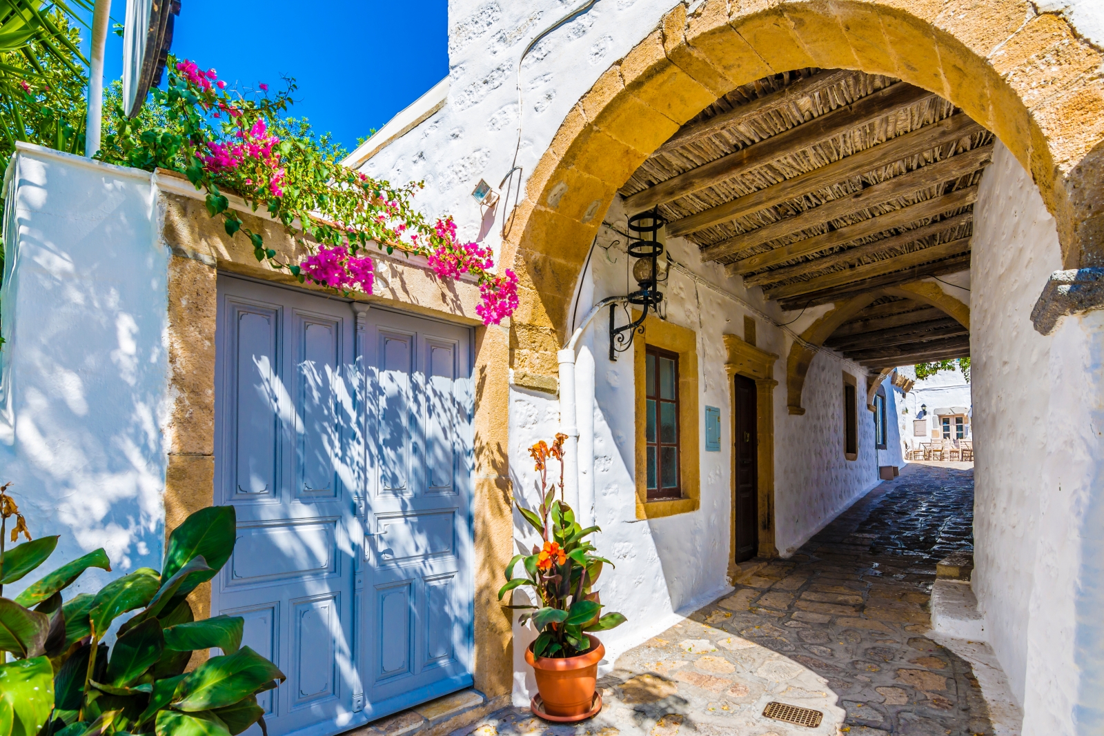 Sunlit alley with arch cover and blue door surrounded by pink flowers