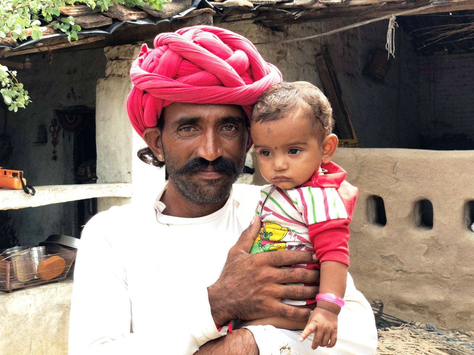 An Indian man with a mustache and wearing a red turban and white shirt holding a baby in India