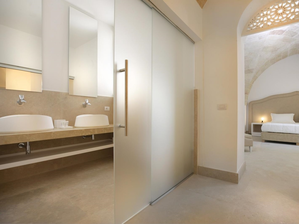 Bedroom with double bed, bedside table, lamp & ensuite bathroom with sinks and mirrors at Masseria dei Papi in Puglia, Italy
