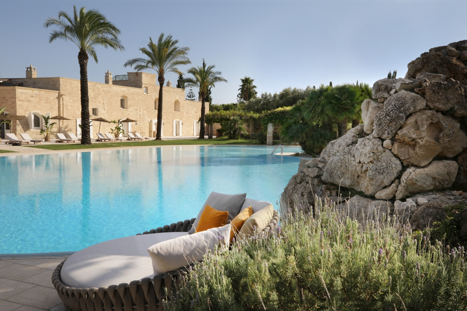 Swimming pool and patio with plants, sofa, sun loungers, umbrellas and palm trees at Masseria dei Papi in Puglia, Italy