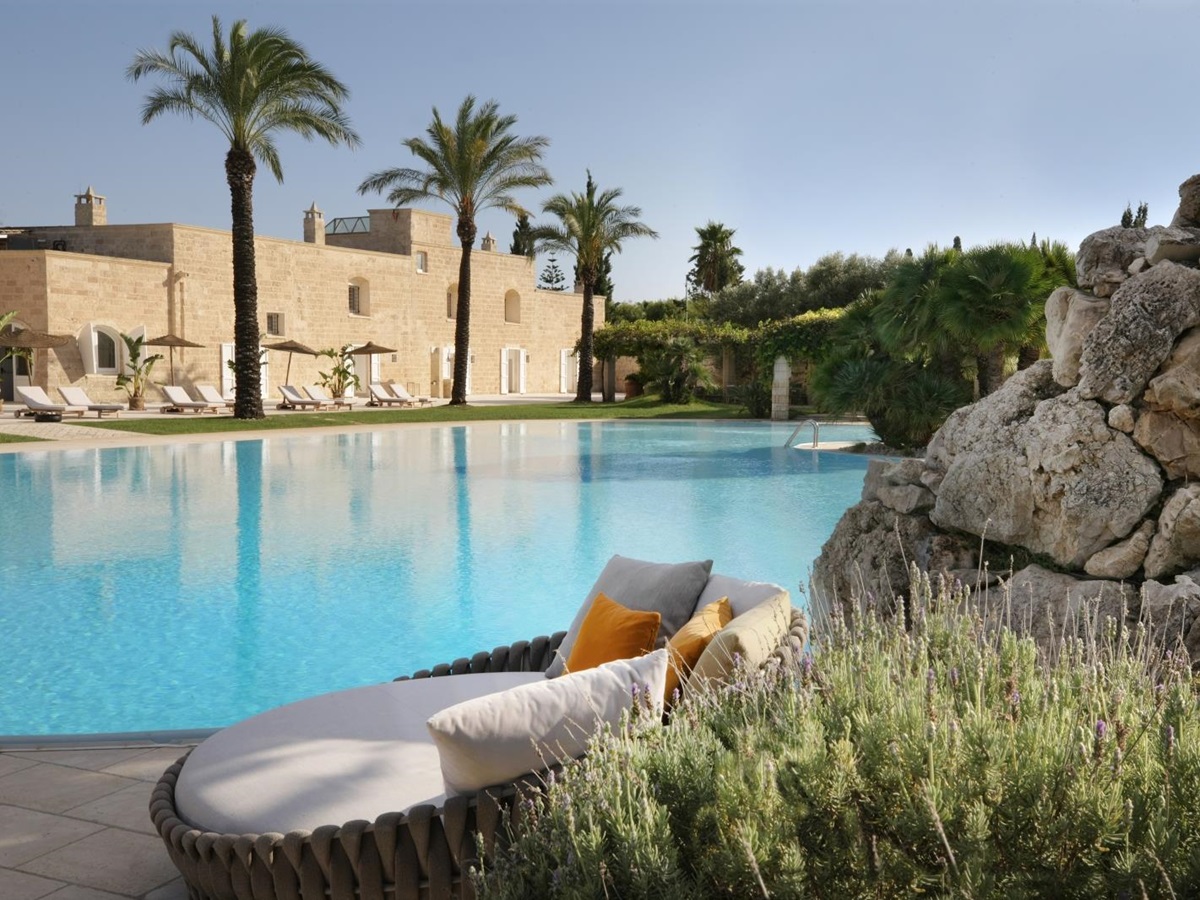 Swimming pool and patio with plants, sofa, sun loungers, umbrellas and palm trees at Masseria dei Papi in Puglia, Italy