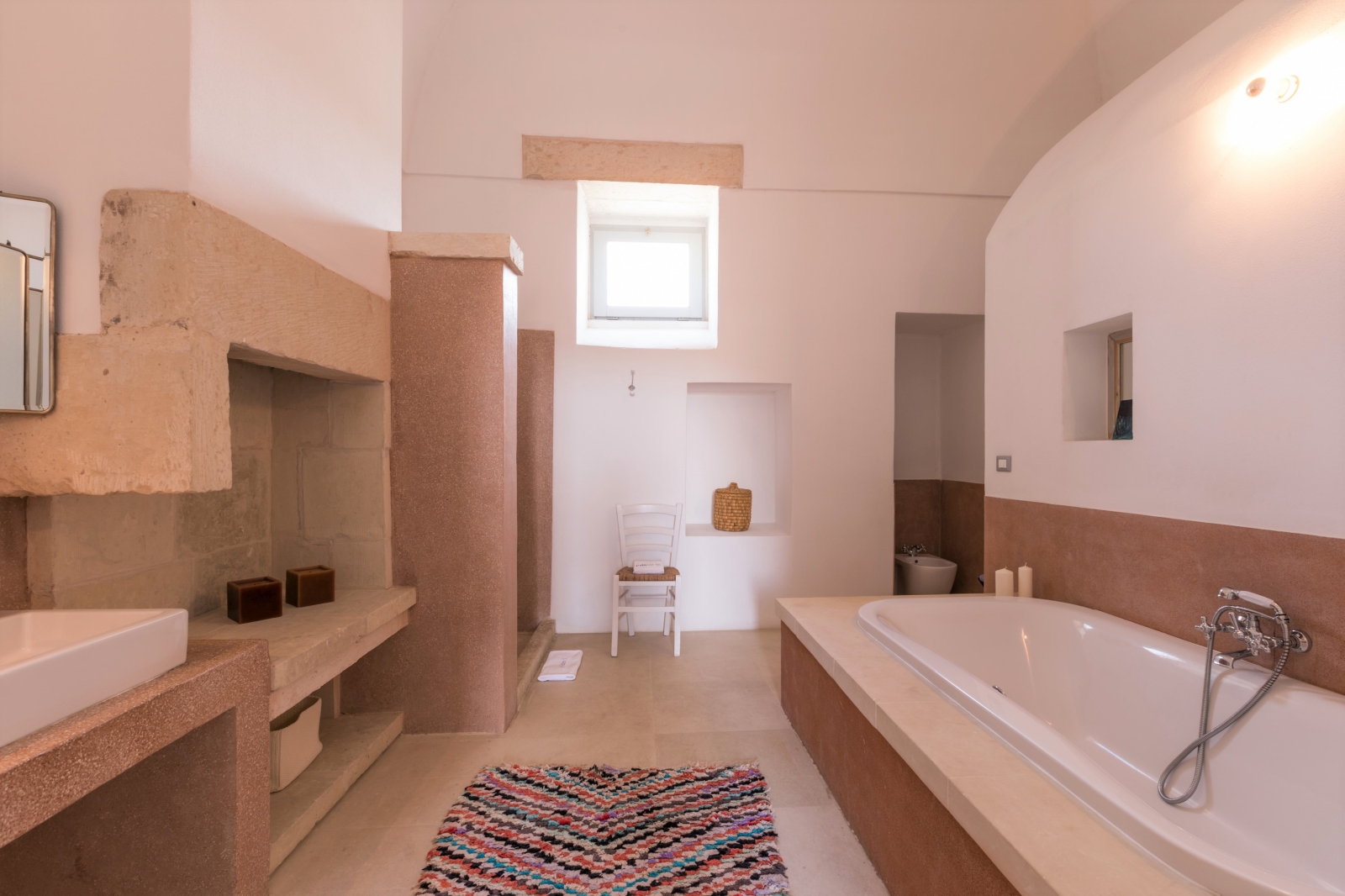 Bathroom with bath, sink, patterned rug, chair and small window at Palazzo del Duca in Puglia, Italy