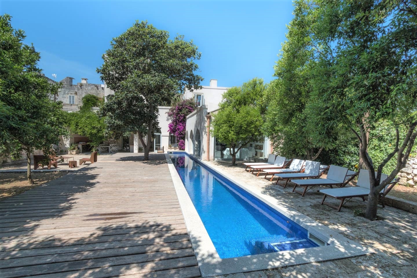 Long swimming pool and decking area with trees, sun loungers, flowers and view of house at Palazzo del Duca in Puglia, Italy