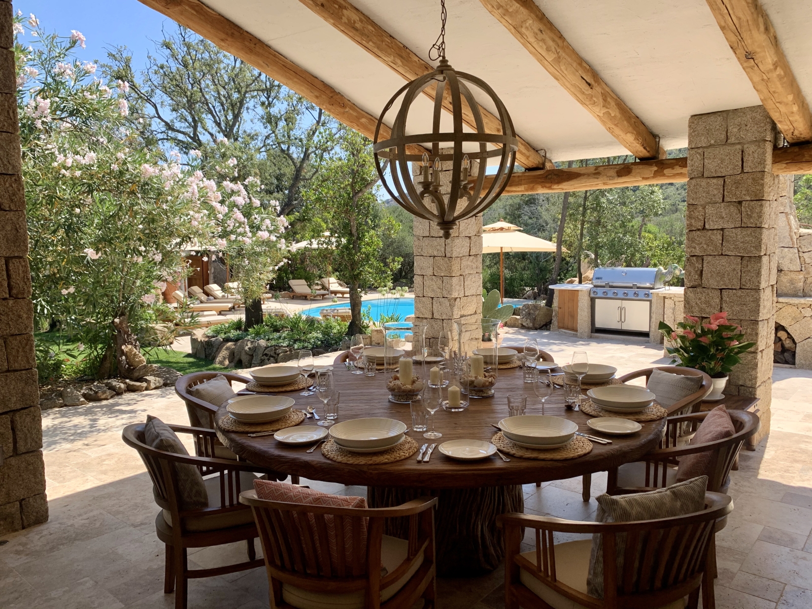 Covered outdoor dining area with table, chairs, hanging light, BBQ and view to pool at Villa Pantaleo on Sardinia, Italy