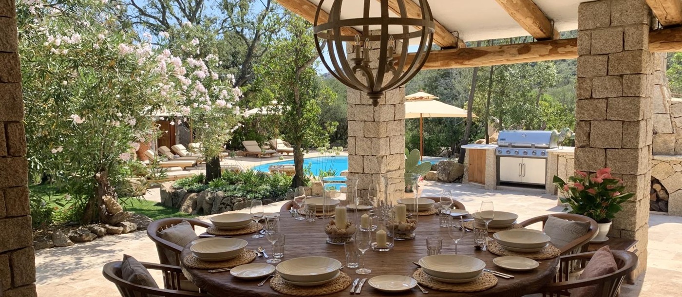 Covered outdoor dining area with table, chairs, hanging light, BBQ and view to pool at Villa Pantaleo on Sardinia, Italy