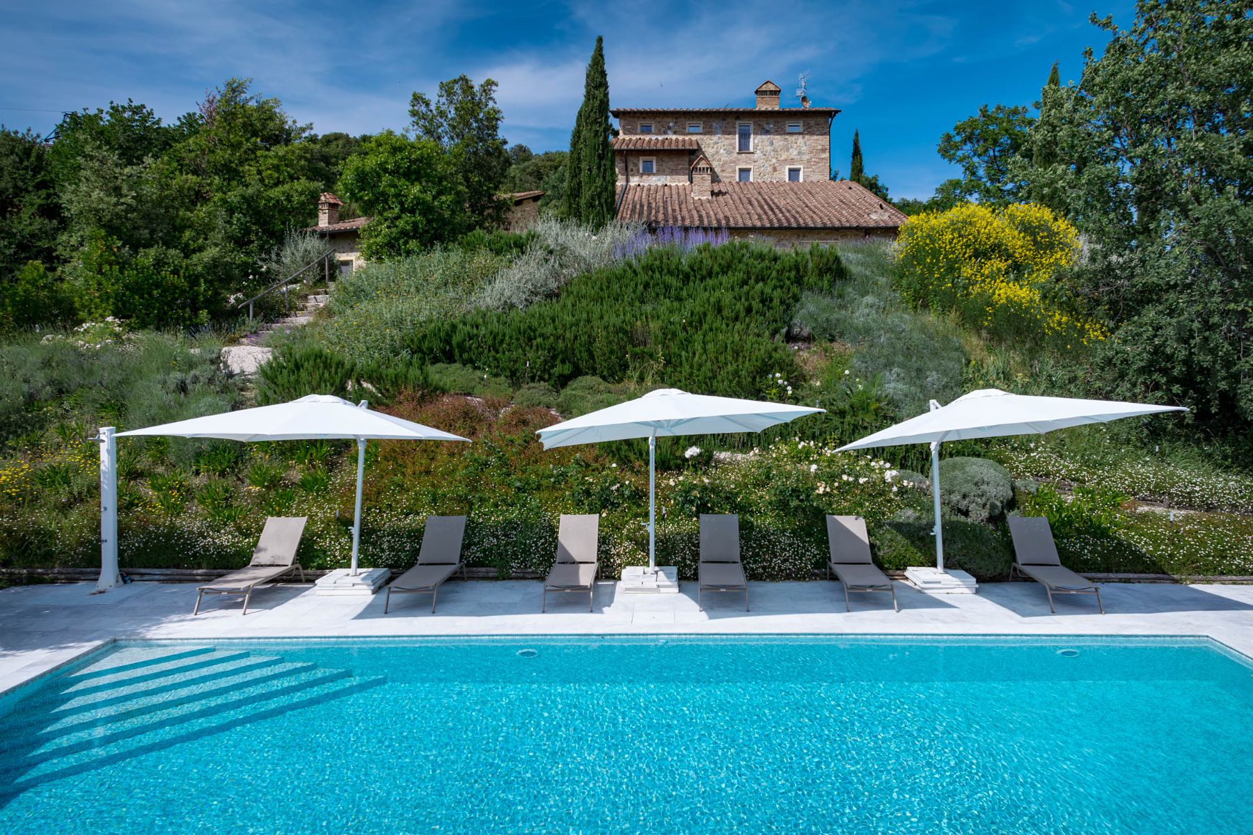 Close view of the outdoor pool featuring sun loungers and umbrellas with view of Villa Il Mugnello in the background in Tuscany