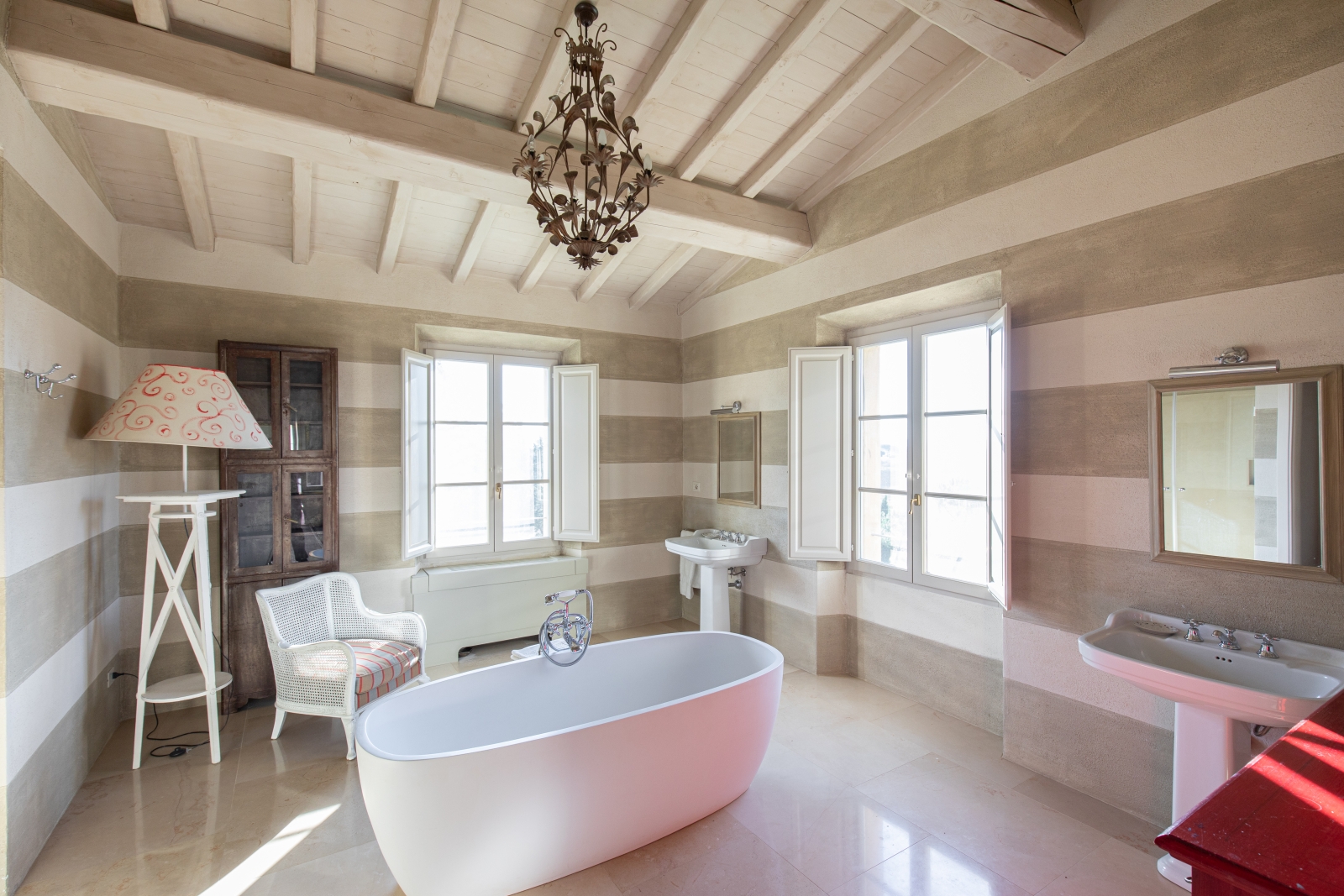 Large bathroom with bath tub, two sinks, armchair, lamp, book case, chandelier and windows at Il Serratone in Tuscany, Italy
