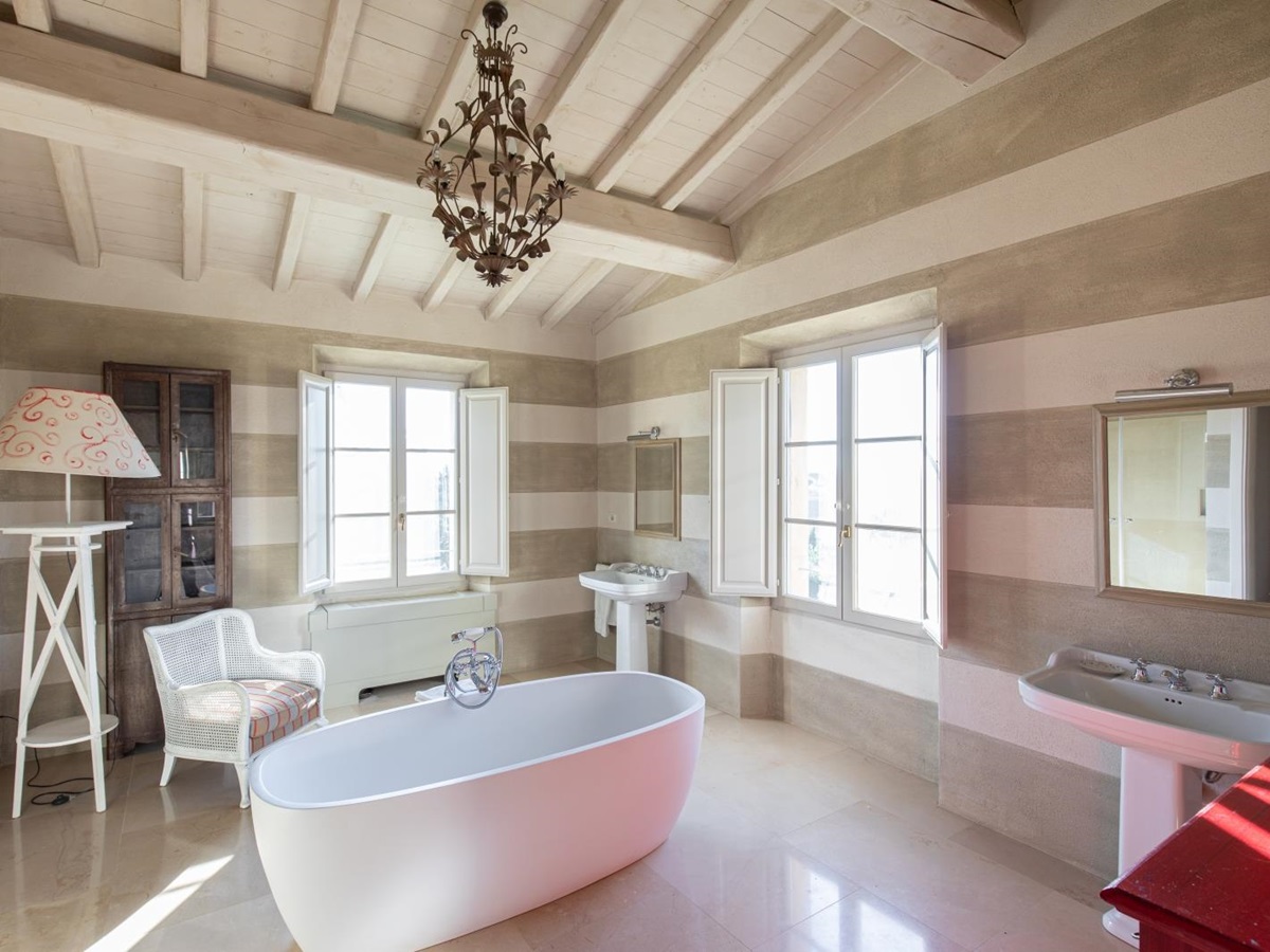 Large bathroom with bath tub, two sinks, armchair, lamp, book case, chandelier and windows at Il Serratone in Tuscany, Italy