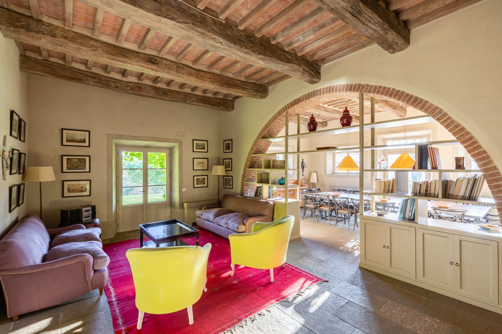 Lounge with sofas, armchairs, table, pictures, shelves and stone archway to dining room at Il Serratone in Tuscany, Italy