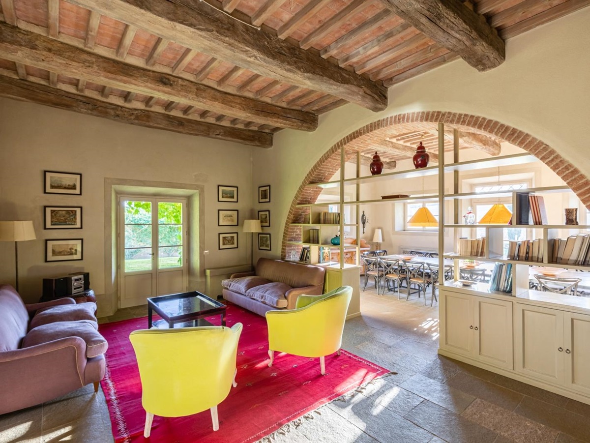 Lounge with sofas, armchairs, table, pictures, shelves and stone archway to dining room at Il Serratone in Tuscany, Italy