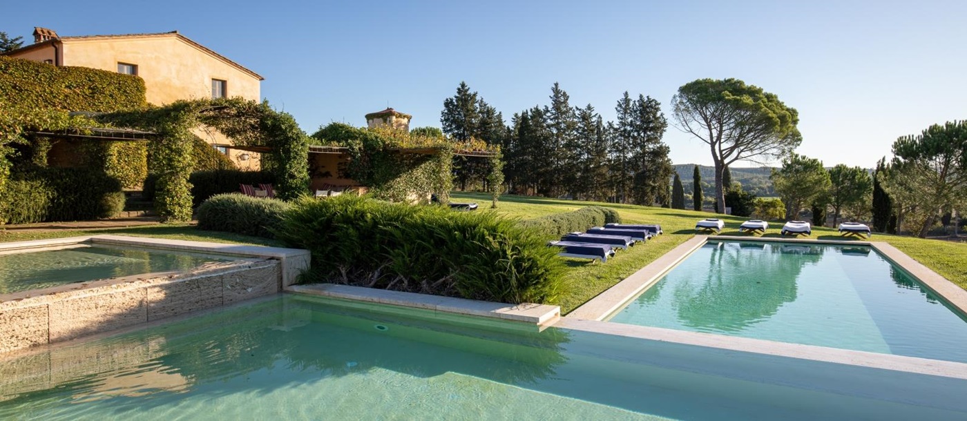 Large swimming pool in garden with sun loungers, grass, hedges & trees at Il Serratone in Tuscany, Italy