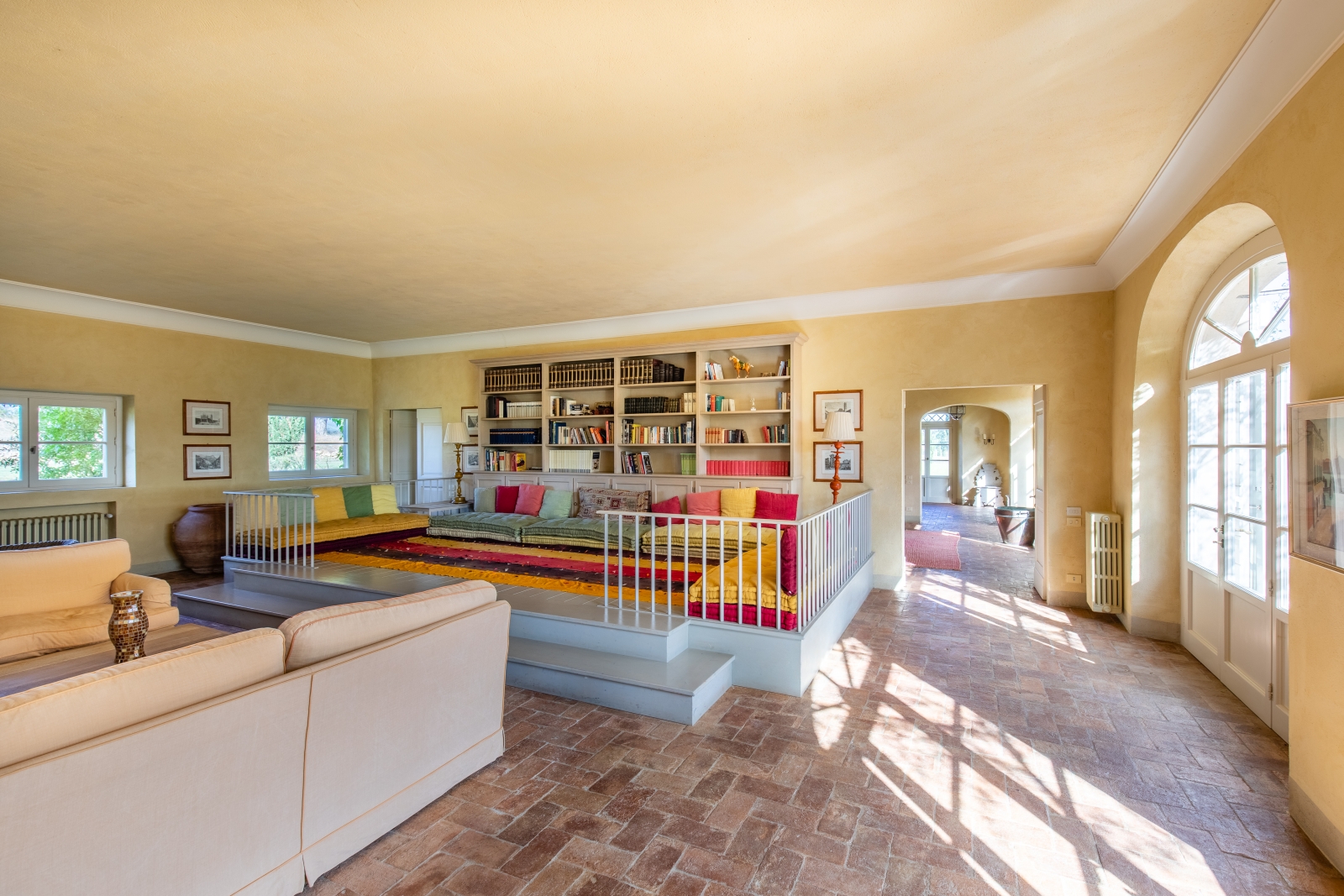 Living room with sofas, colourful elevated sitting area, bookshelves, lamps and French doors at La Vigna in Tuscany, Italy