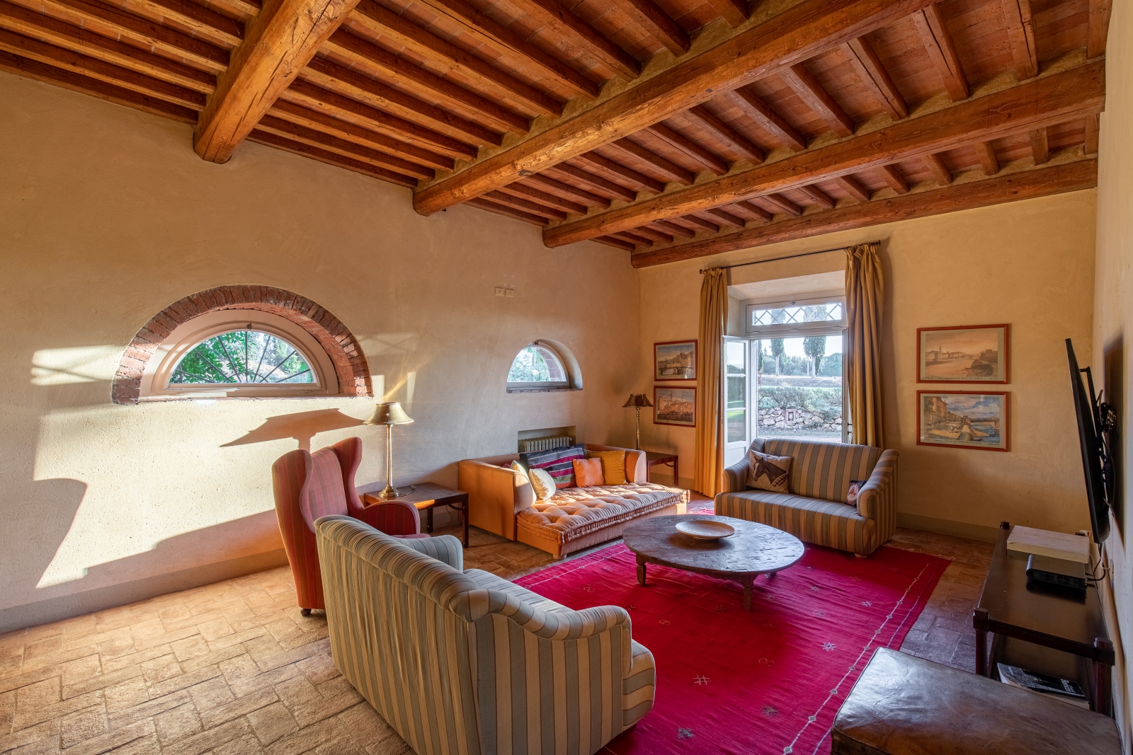 Living room with pink rug, coffee table, sofas, armchair, TV and French doors to garden at La Vigna in Tuscany, Italy