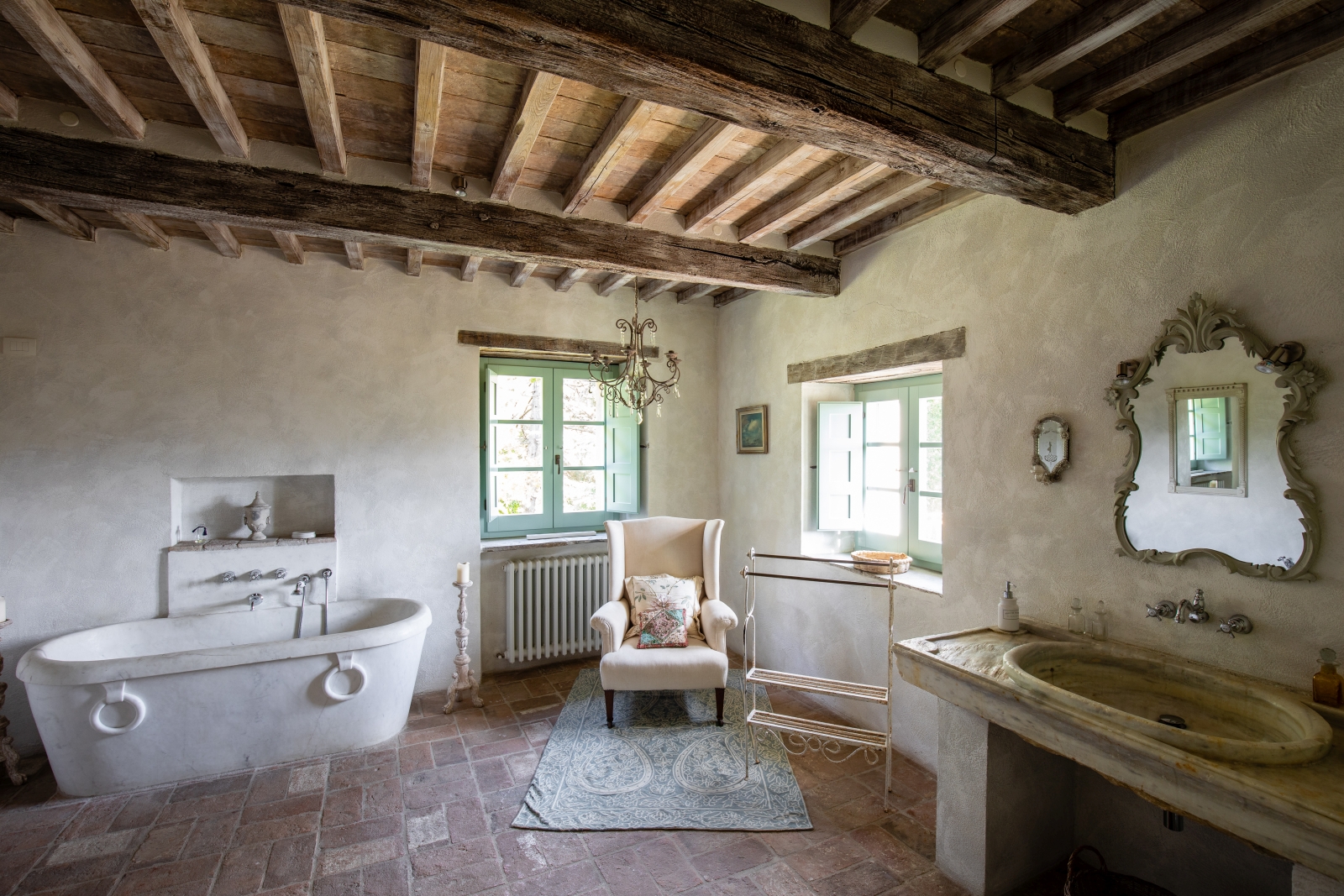 Bathroom with bath, candle, armchair, windows, chandelier, sink, mirror and ceiling beams at Tenuta Cantata in Tuscany, Italy