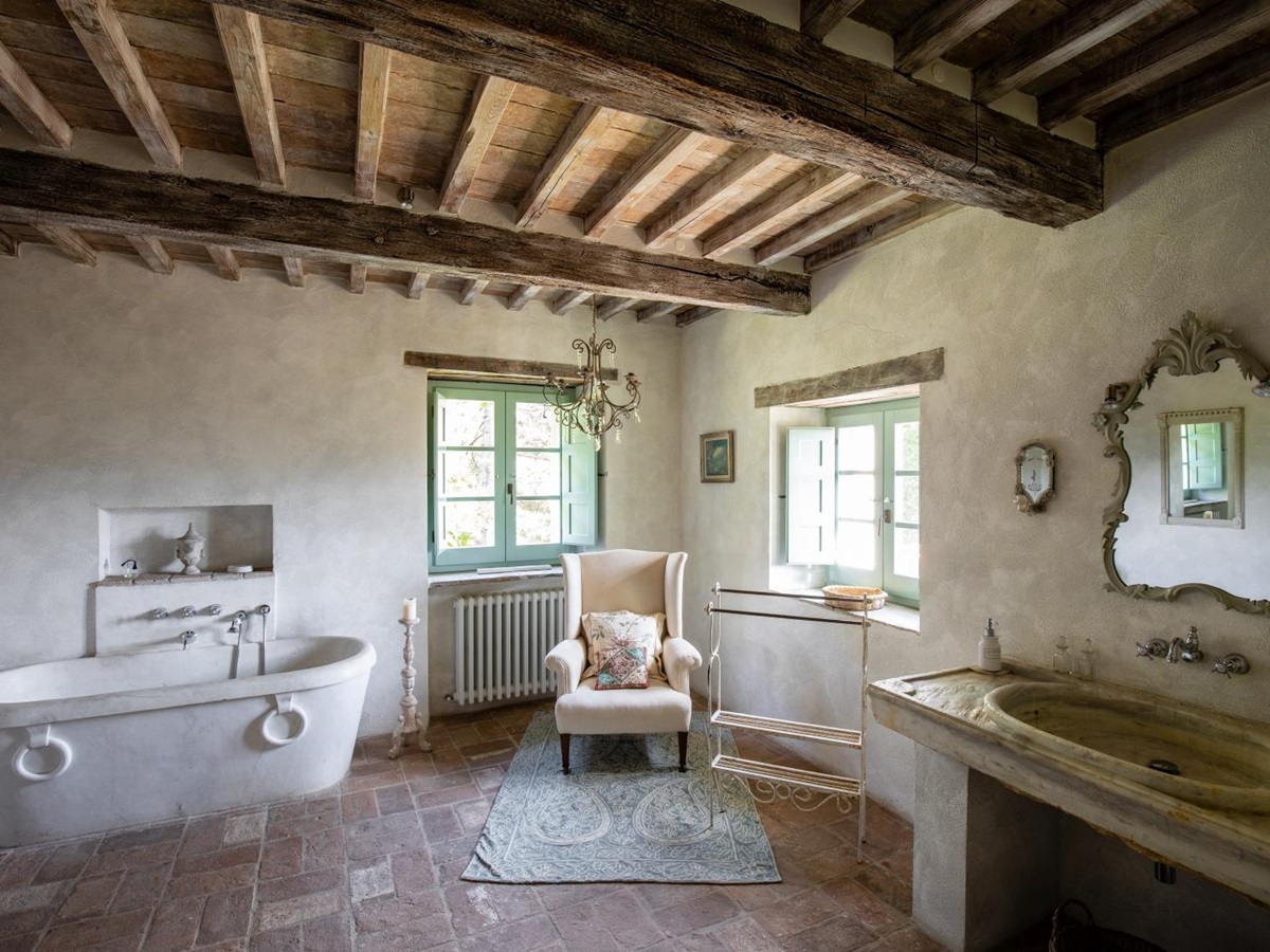 Bathroom with bath, candle, armchair, windows, chandelier, sink, mirror and ceiling beams at Tenuta Cantata in Tuscany, Italy
