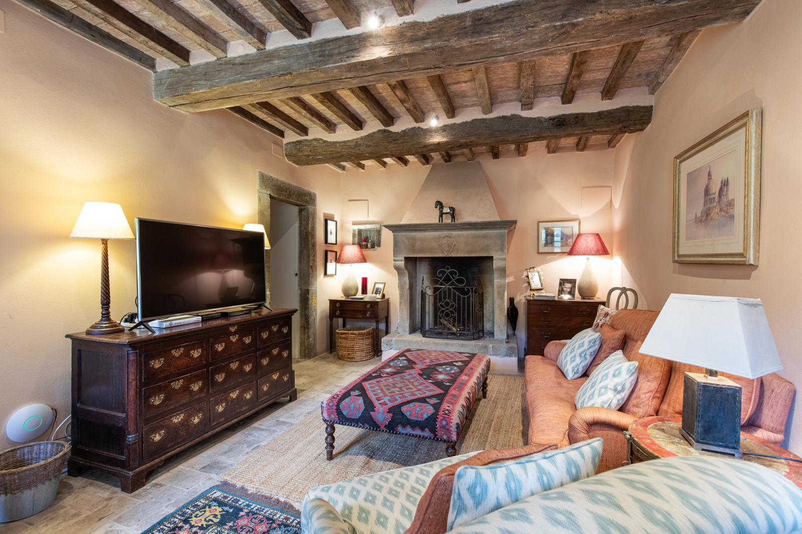 Living room with TV, sofa, armchair, footstool, artwork, cabinets, lamps and fireplace at Tenuta Cantata in Tuscany, Italy