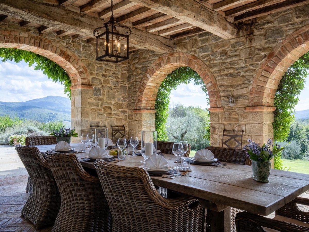 Outdoor dining area with loggia, set table, chairs, light, flowers and countryside view at Tenuta Cantata in Tuscany, Italy