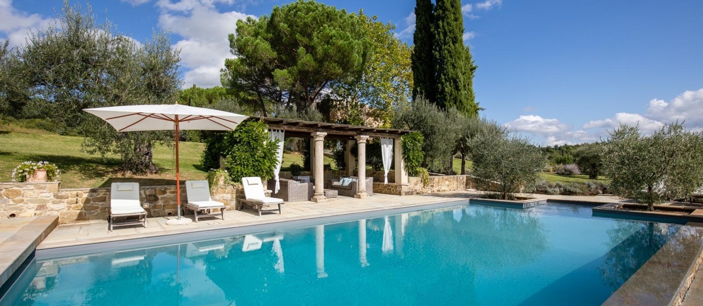 Pool with patio area, sun loungers, covered lounge area, trees and view of garden at Tenuta Cantata in Tuscany, Italy