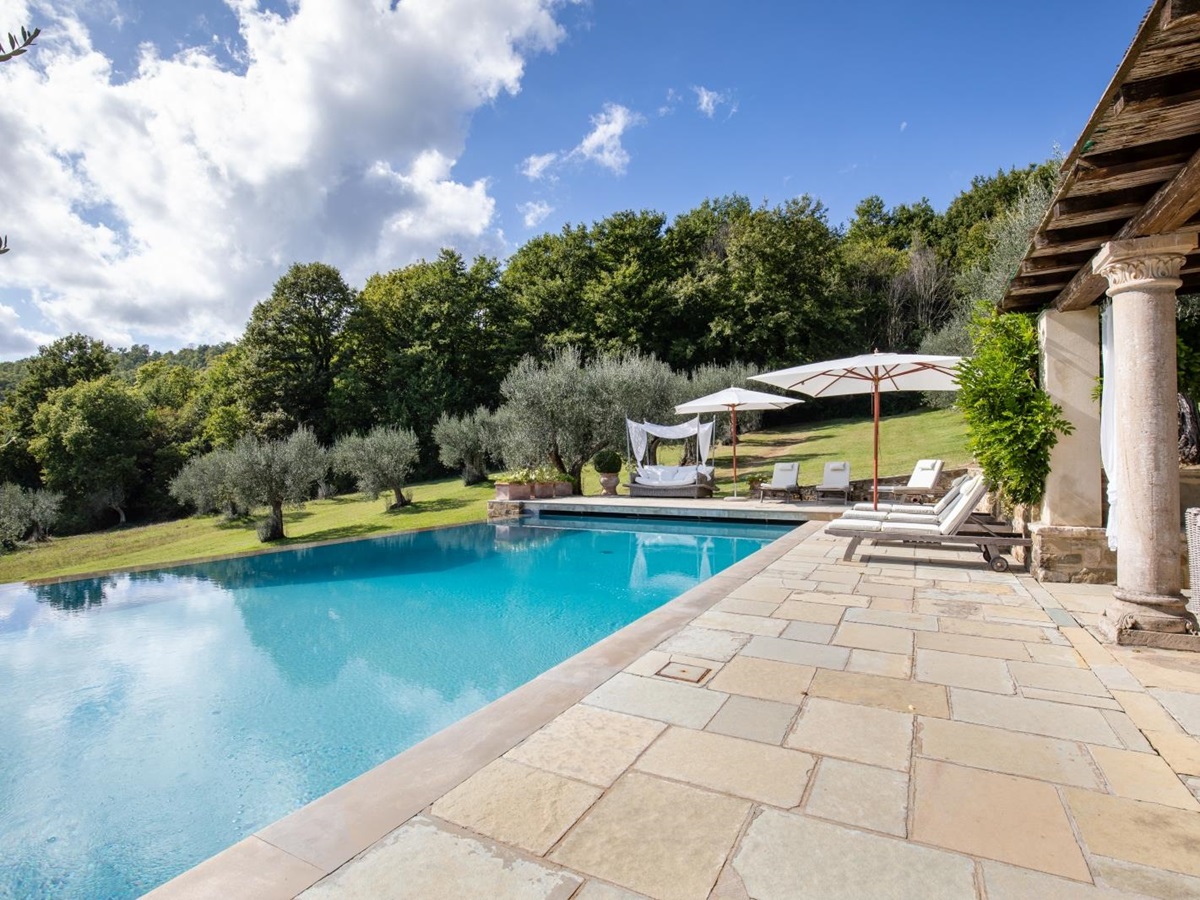 Pool area with patio, stone pillars, sun loungers, umbrellas, day bed and view of garden at Tenuta Cantata in Tuscany, Italy
