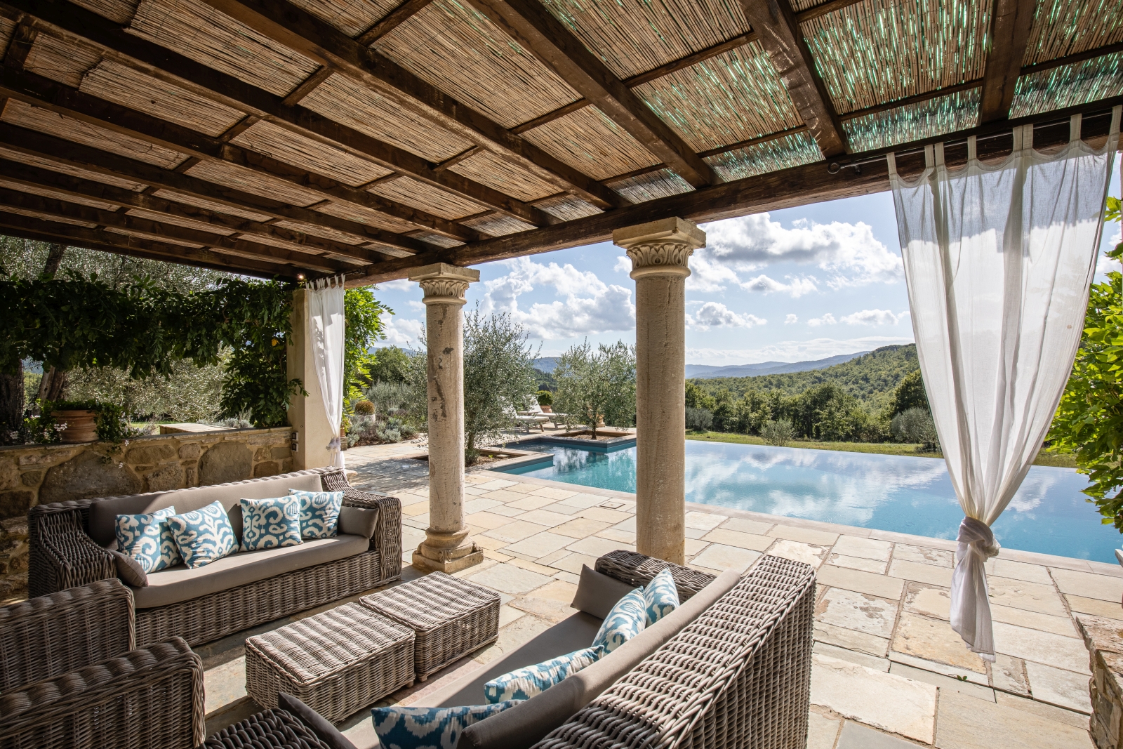 Covered area by pool with sofas, coffee tables, curtains, stone pillars and garden view at Tenuta Cantata in Tuscany, Italy