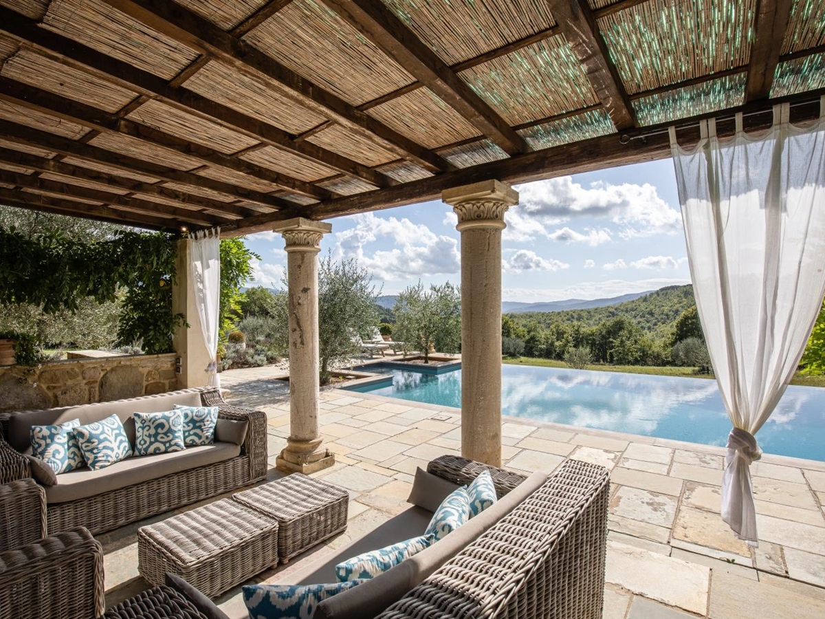 Covered area by pool with sofas, coffee tables, curtains, stone pillars and garden view at Tenuta Cantata in Tuscany, Italy