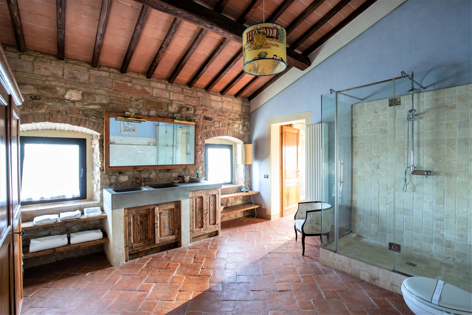Bathroom with sinks, large mirror, armchair, large shower, toilet, stone wall and windows at Villa Giuliana in Tuscany, Italy