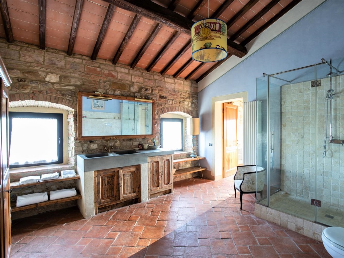 Bathroom with sinks, large mirror, armchair, large shower, toilet, stone wall and windows at Villa Giuliana in Tuscany, Italy