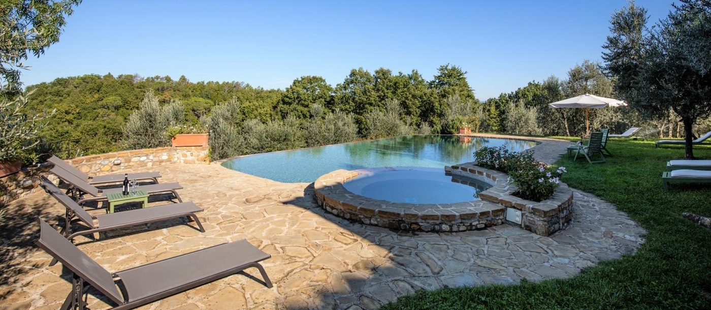 Patio next to pool with sun loungers, table, wine, umbrella and countryside view at Villa Giuliana in Tuscany, Italy