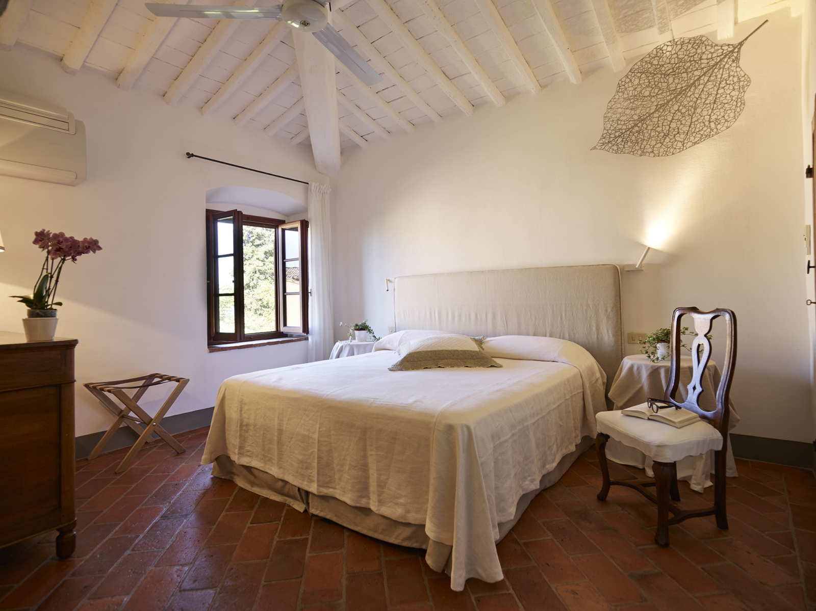 Bedroom with double bed, bedside tables, plants, flowers, chair and window at Villa La Castellana in Tuscany, Italy