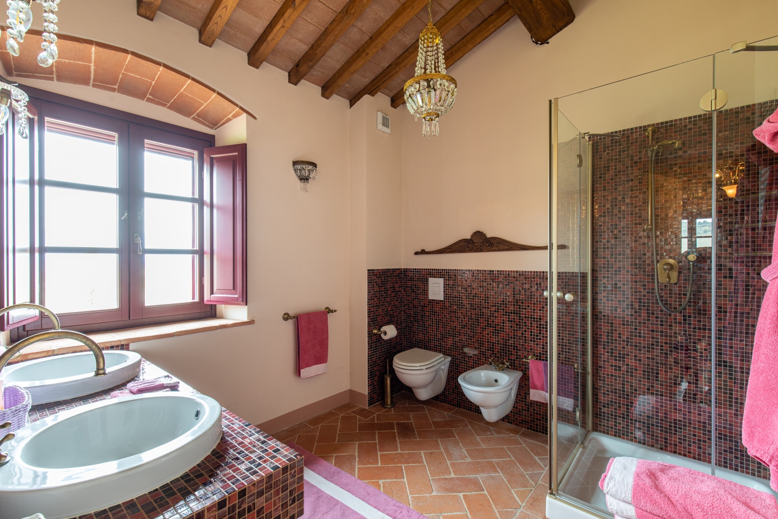 Bathroom with red and black tiles, sinks, toilet, bidet, chandelier, window and shower at Villa Papavero in Tuscany, Italy