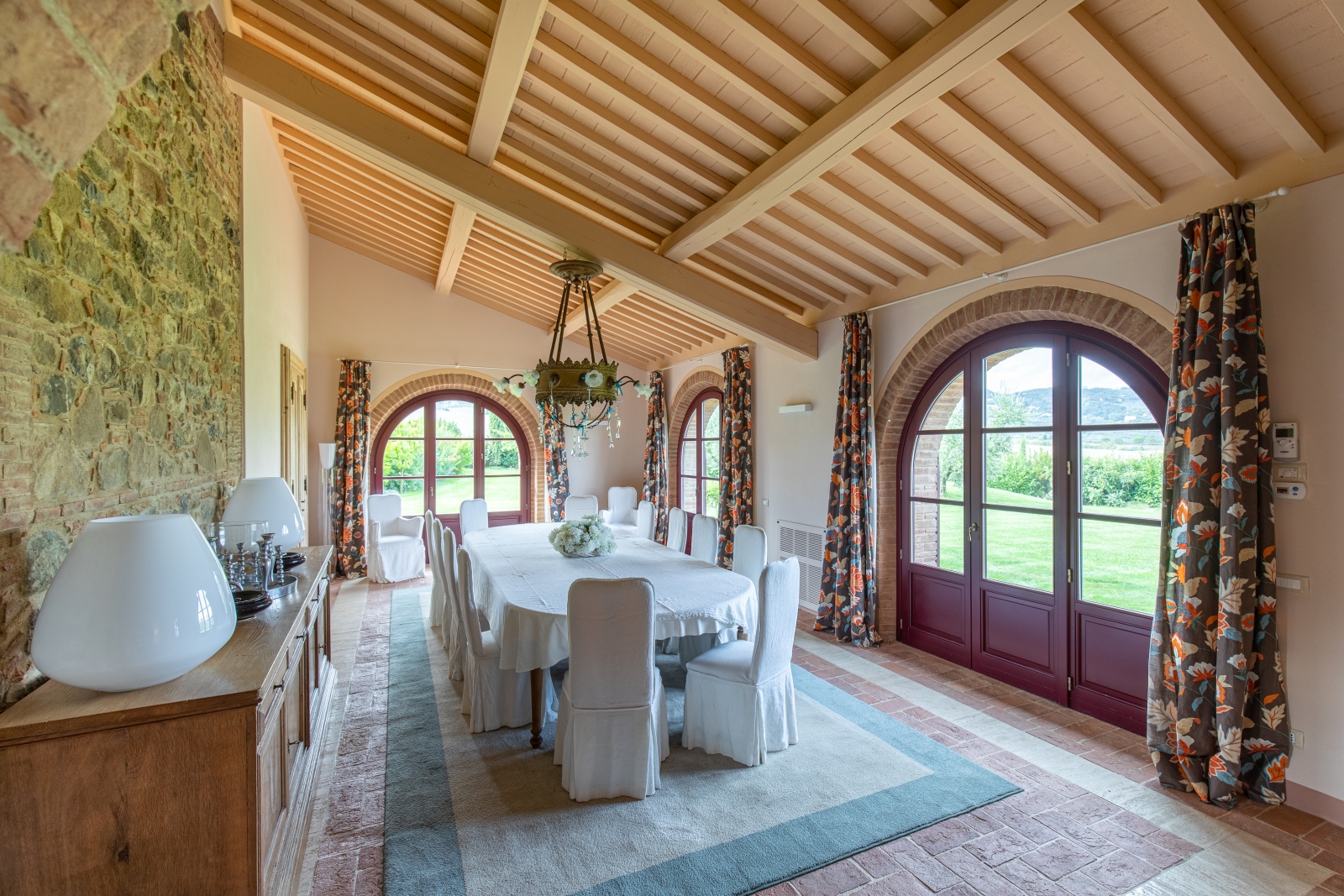 Dining room with long table, chairs, rug, cabinet, lamps, chandelier and doors to garden at Villa Papavero in Tuscany, Italy
