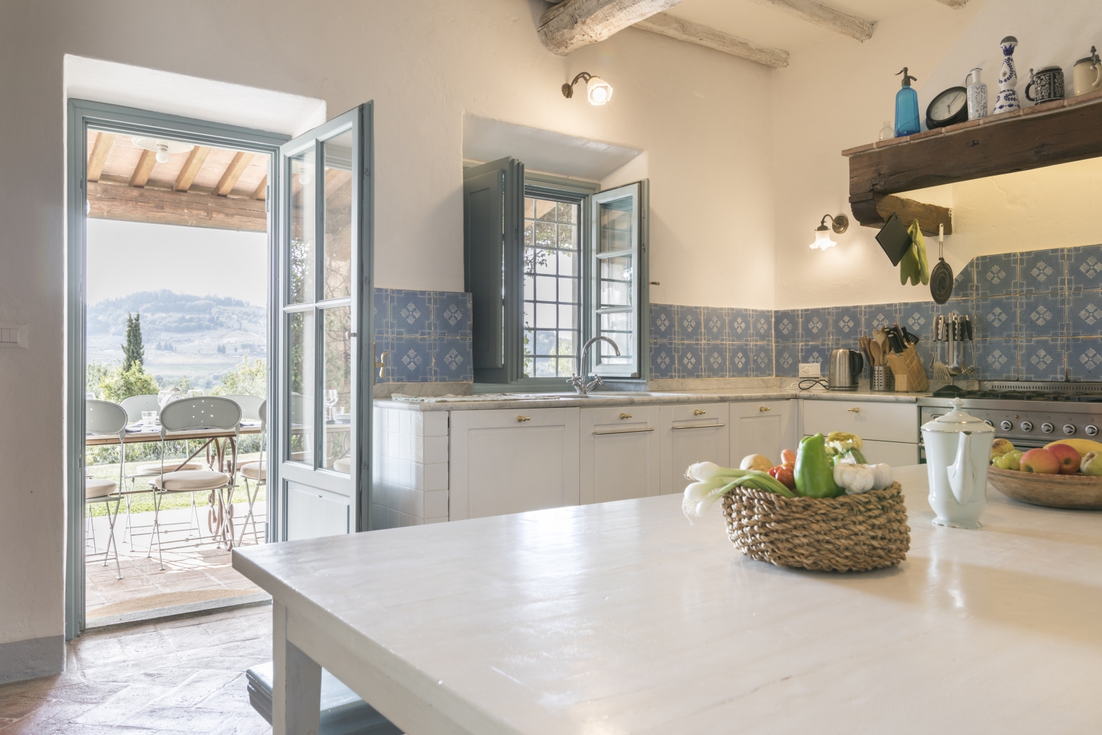 Kitchen with island, blue tiles, shelves, fresh food and French doors to terrace at Villa San Casciano in Tuscany, Italy