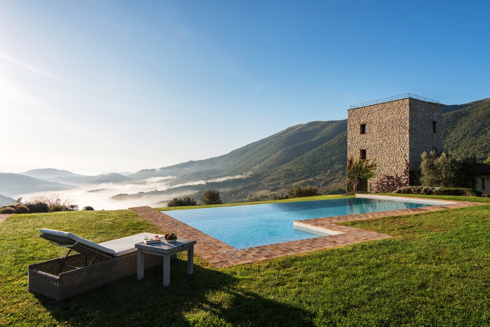 Pool with sun lounger, coffee table, fruit, green grass and view of mountains and villa at Bel Canto in Umbria, Italy