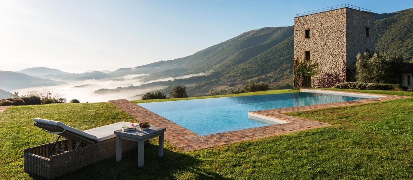 Pool with sun lounger, coffee table, fruit, green grass and view of mountains and villa at Bel Canto in Umbria, Italy