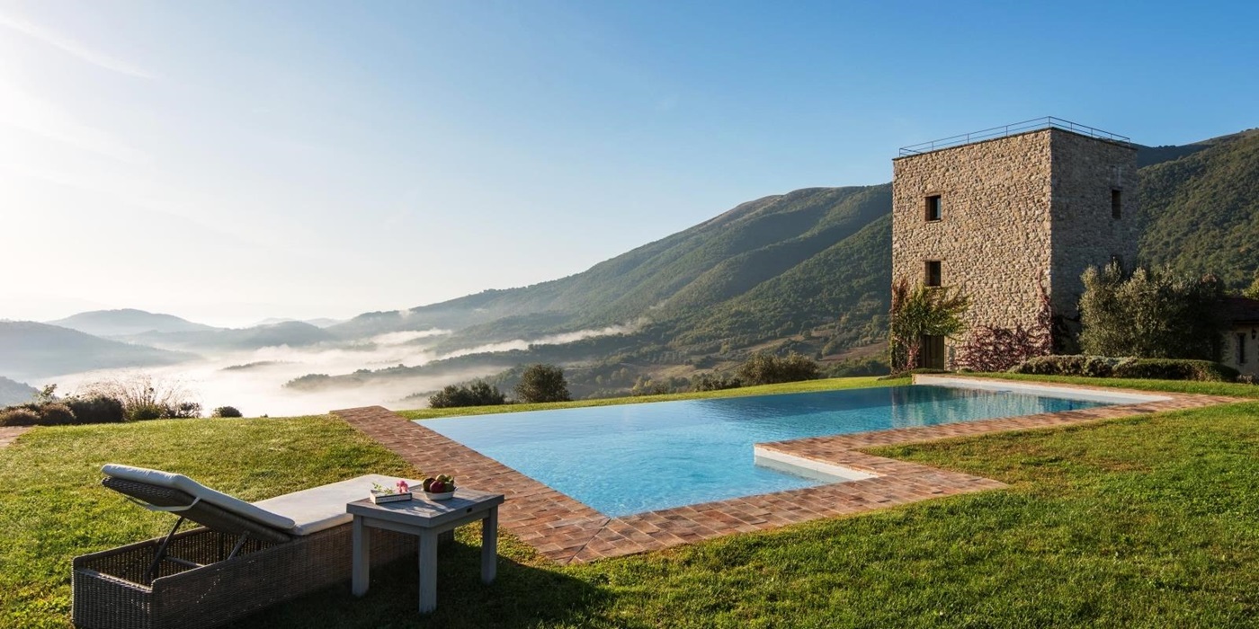Pool with sun lounger, coffee table, fruit, green grass and view of mountains and villa at Bel Canto in Umbria, Italy