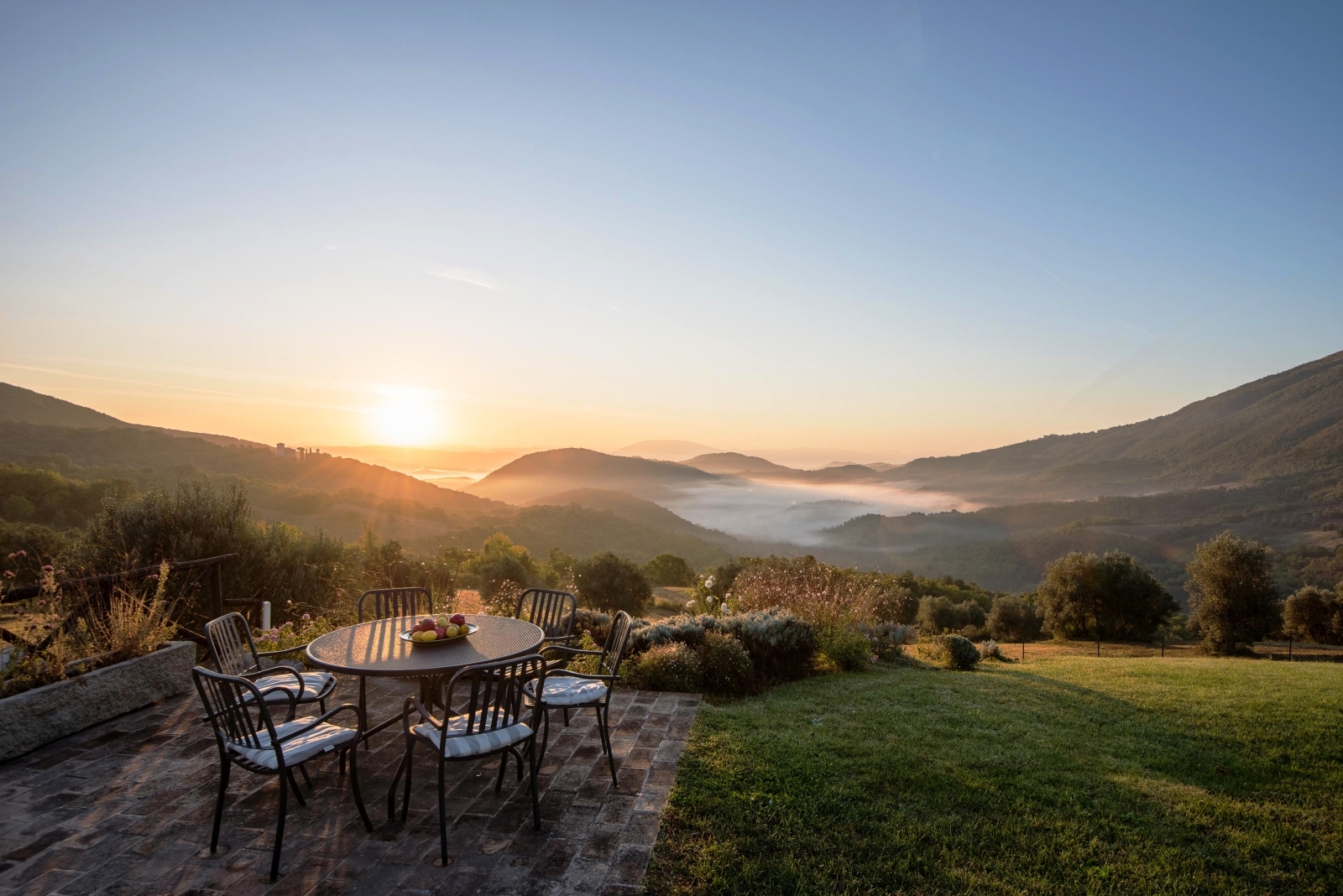 Patio with table, chairs, fresh fruit, grass, plants, trees and view of mountains and sunset at Bel Canto in Umbria, Italy