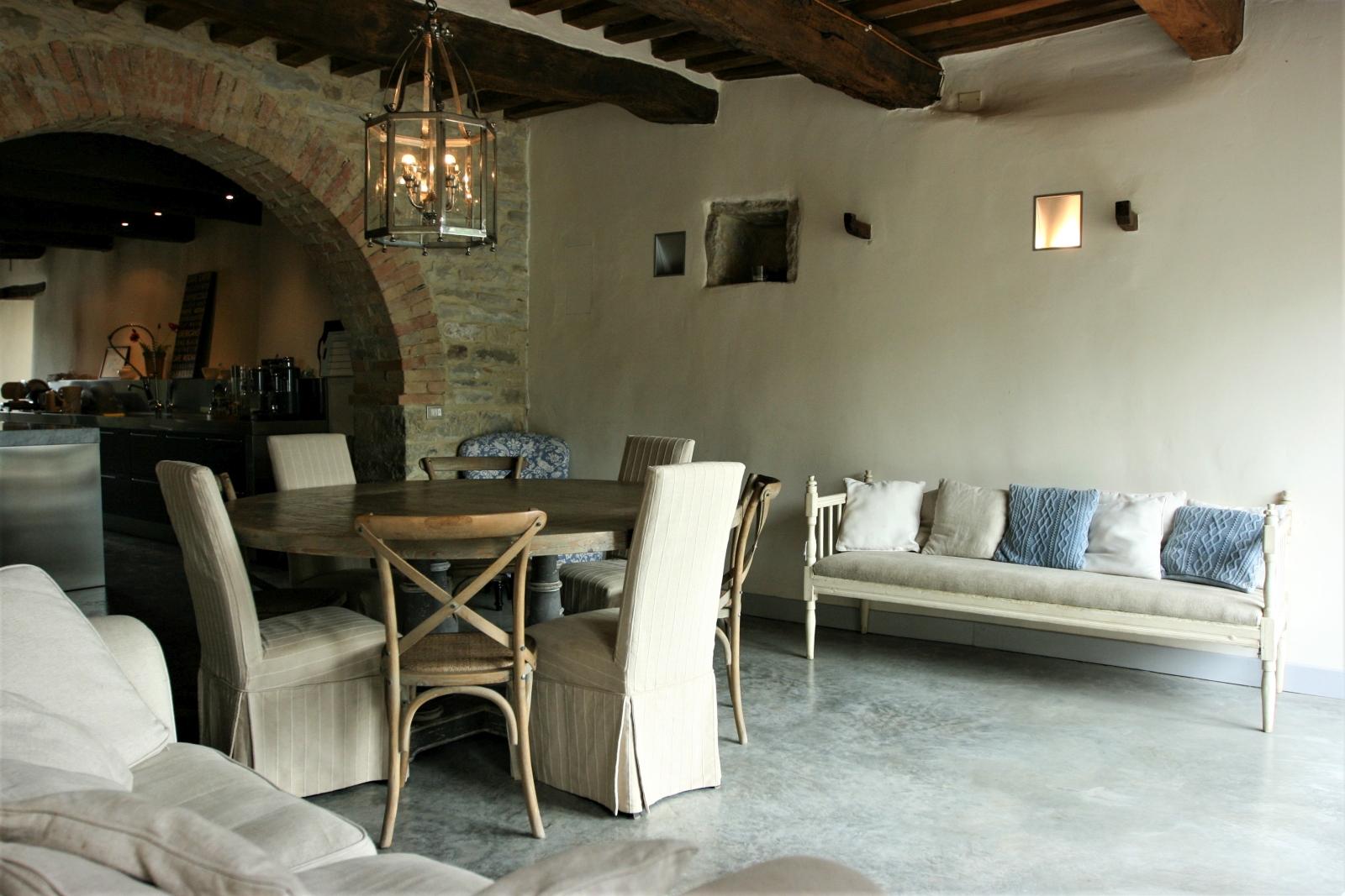 Dining area next to kitchen with round table, chairs, sofas and hanging light at La Spiga in Umbria, Italy