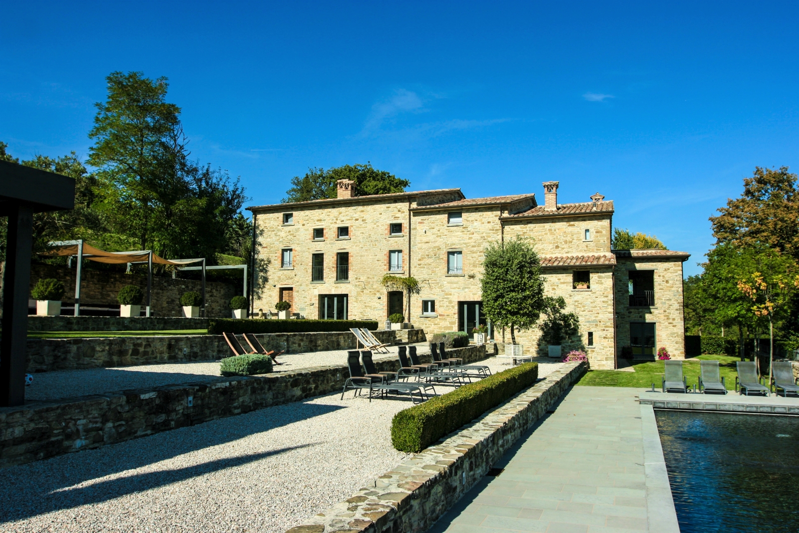 Back of villa with swimming pool, sun loungers, deck chairs, trees and plants at La Spiga in Umbria, Italy