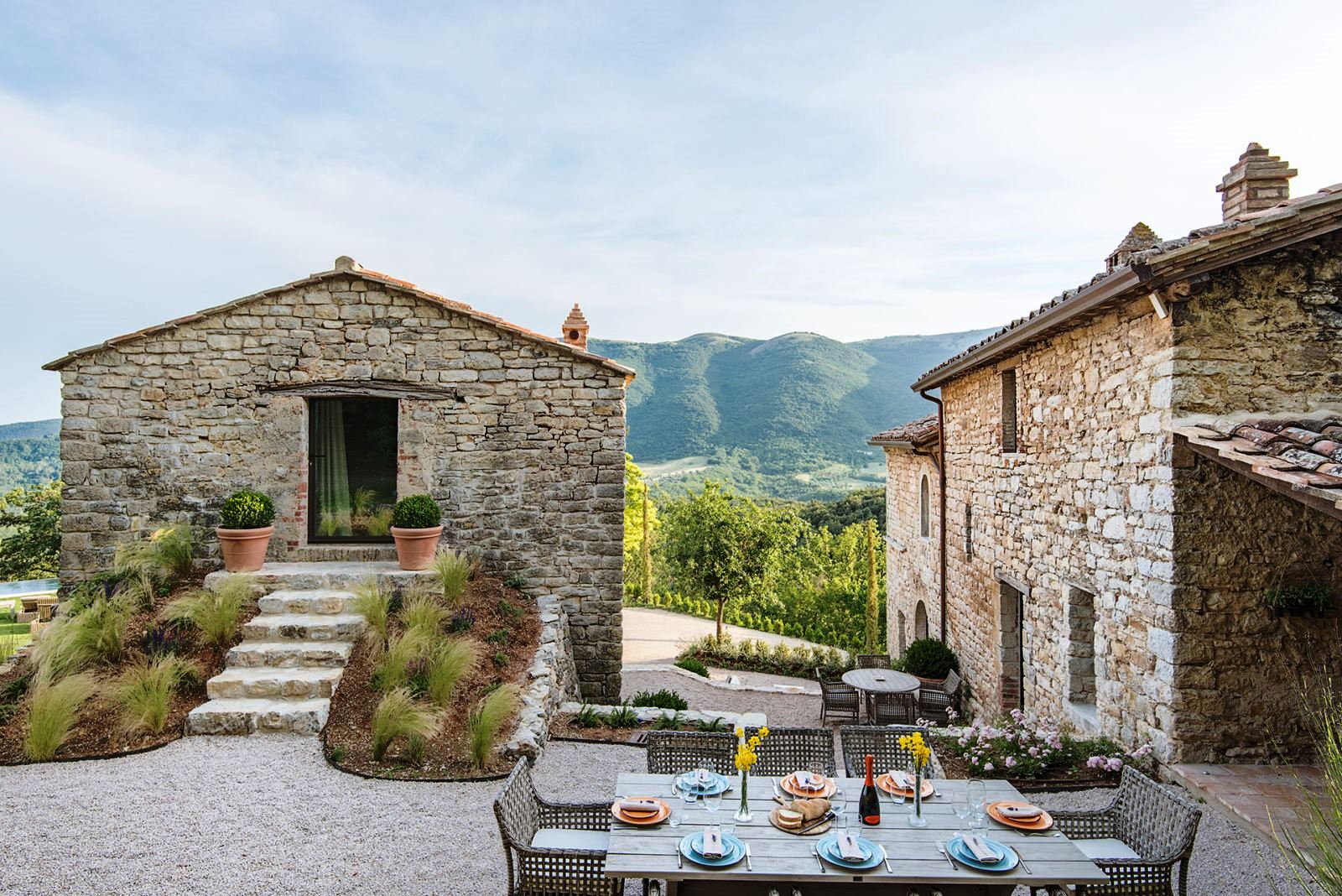 Outdoor dining table with house and annex behind and view of mountains at Villa Piuma in Umbria, Italy