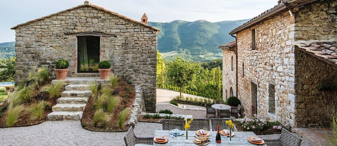 Outdoor dining table with house and annex behind and view of mountains at Villa Piuma in Umbria, Italy