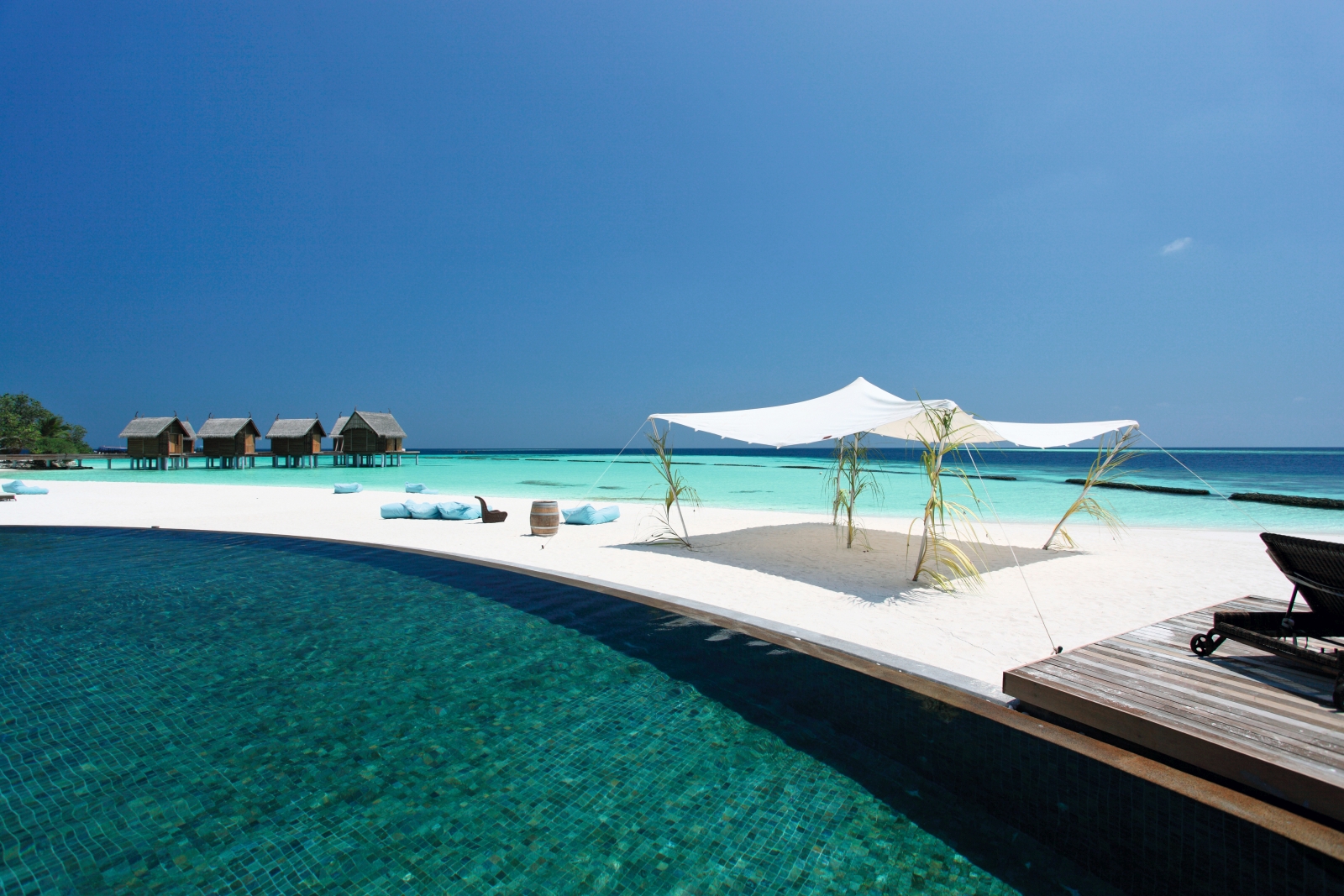 White sand beach with sun sail and loungers separating the turquoise waters of the Indian Ocean and the pool of luxury resort Constance Moofushi in the Maldives