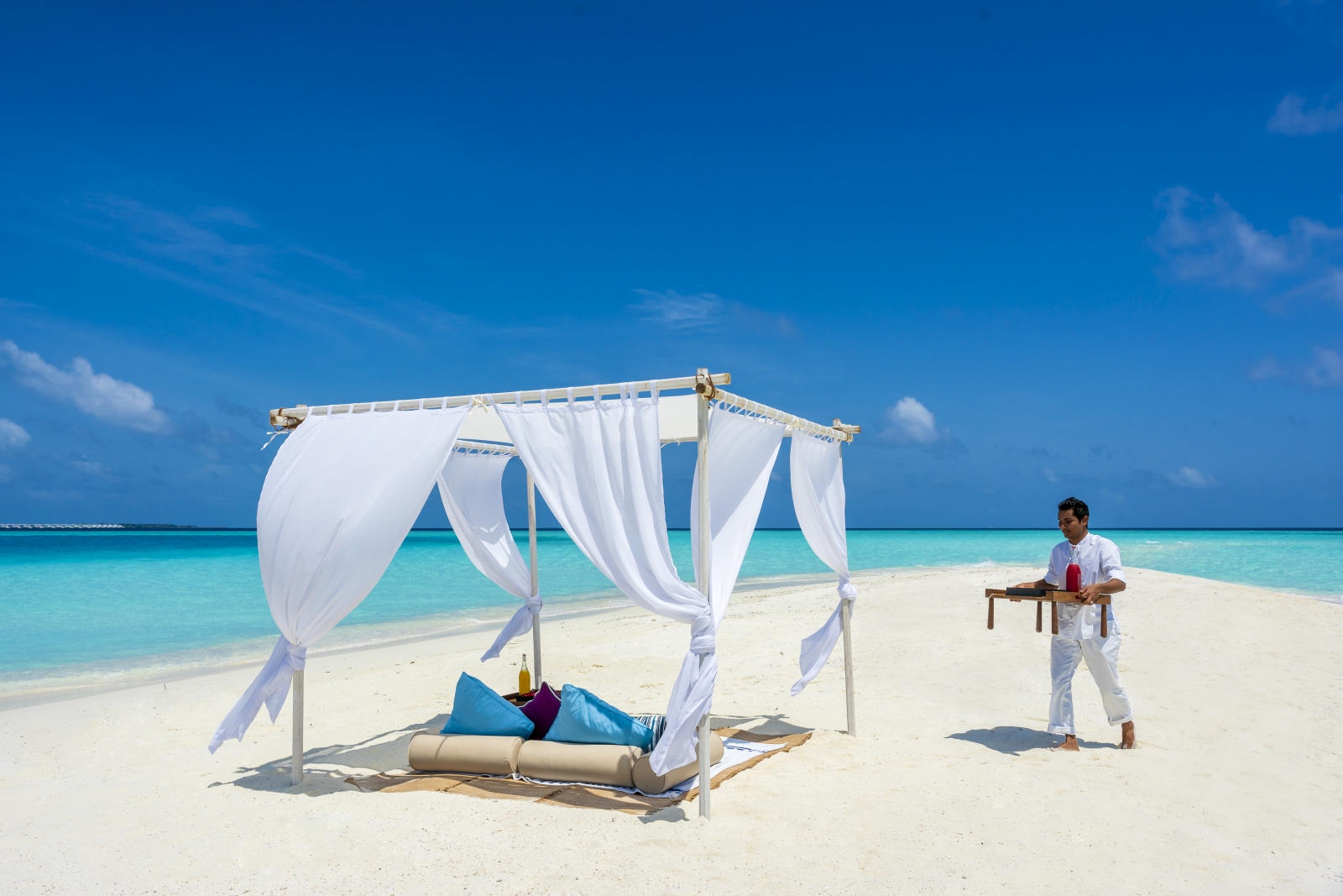 A staff member bringing fresh drinks to a private beach cabana, set on the white sand beach of Milaidhoo surrounded by the tuquoise waters of the Indian Ocean