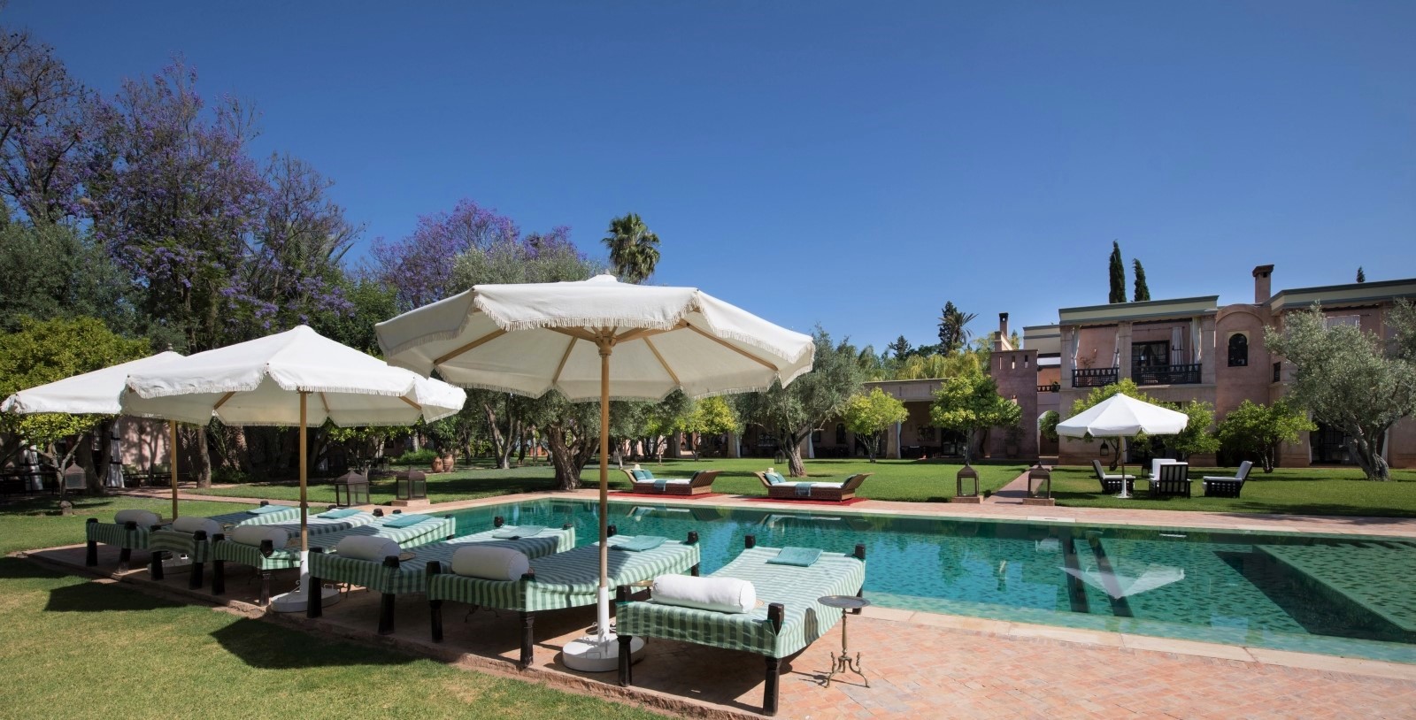 Garden and pool area with sun loungers, umbrellas, lanterns and purple trees at Ezzahra in Marrakech, Morocco