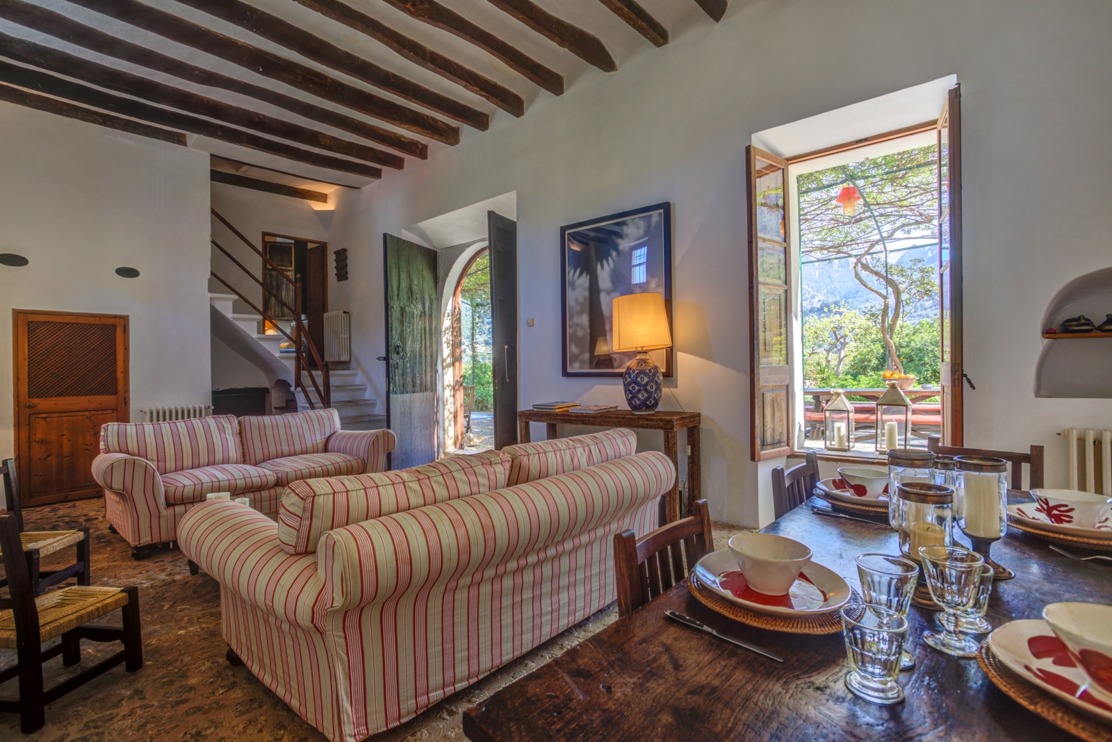 Lounge with dining table, chairs, crockery, two sofas, staircase, window and French doors at Can Canals in Mallorca, Spain