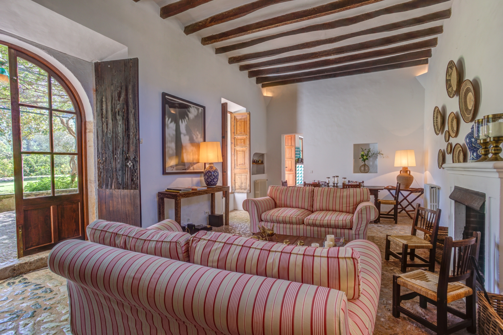 Lounge with sofas, chairs, fireplace, lamps, console table, large window and French doors at Can Canals in Mallorca, Spain