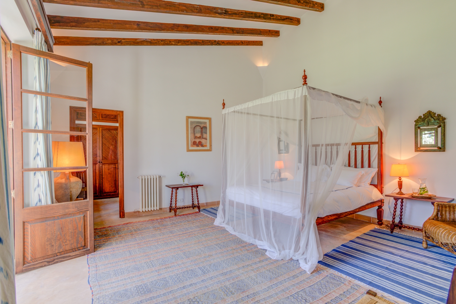 Master bedroom with four-poster double bed, tables, lamps, mirror, painting and French doors at Can Canals in Mallorca, Spain