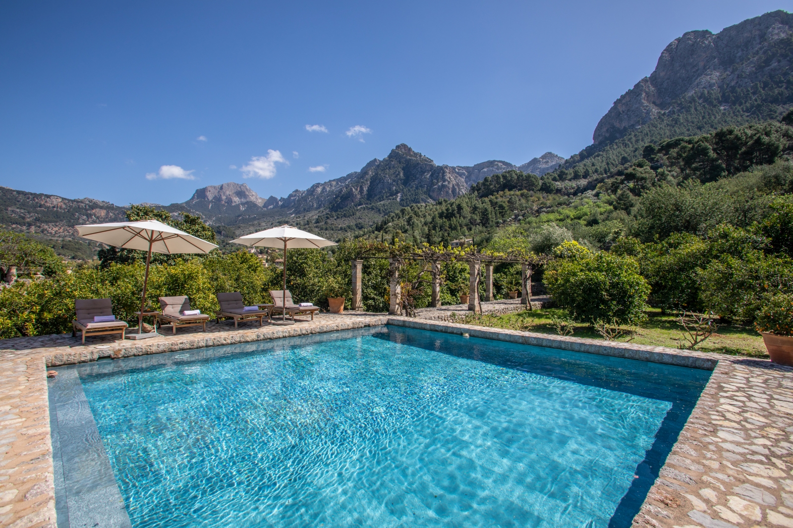 Pool with four sun loungers, two umbrellas, arbour with stone pillars and view of mountains at Can Canals in Mallorca, Spain