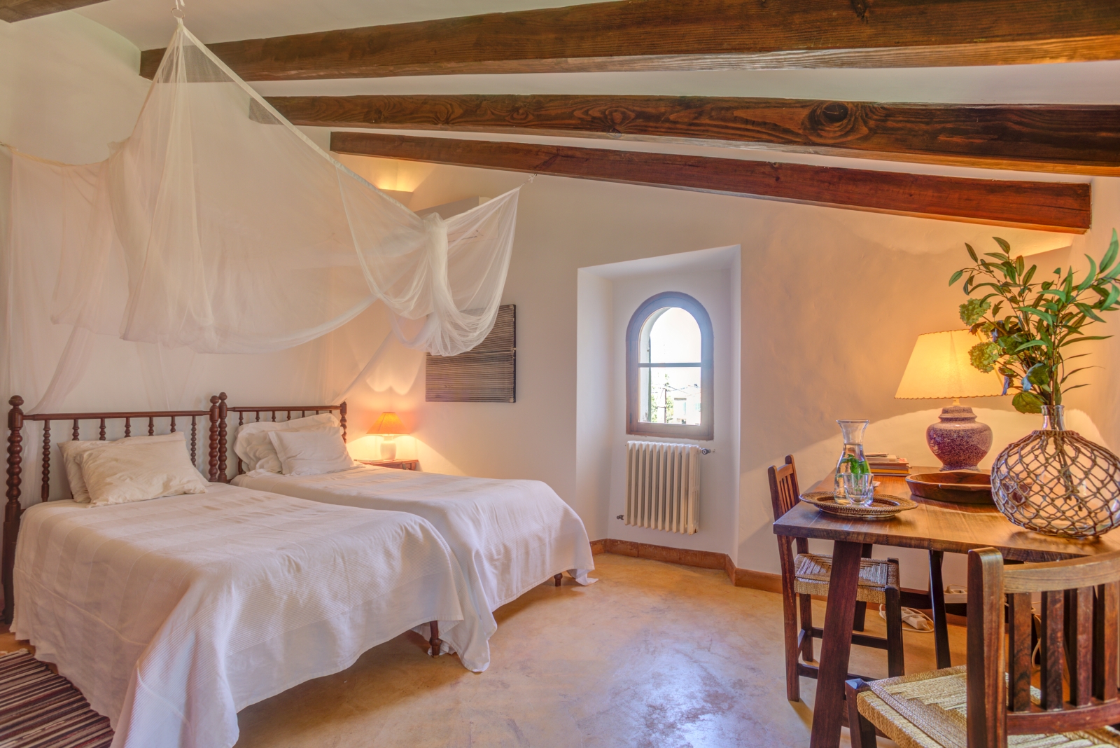 Twin bedroom with net draping, table, chairs, lamp, small window and ceiling beams at Can Canals in Mallorca, Spain